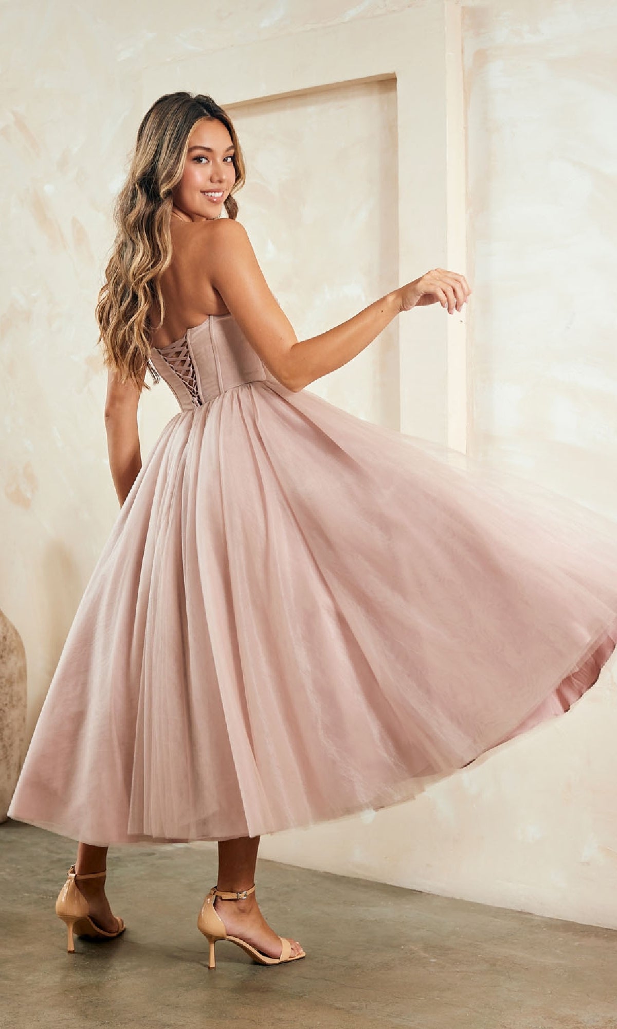 A woman twirls and smiles indoors on a concrete floor wearing the Strapless Pink Semi-Formal Dress: Amelia Couture, a tea-length dress with a lace-up back.